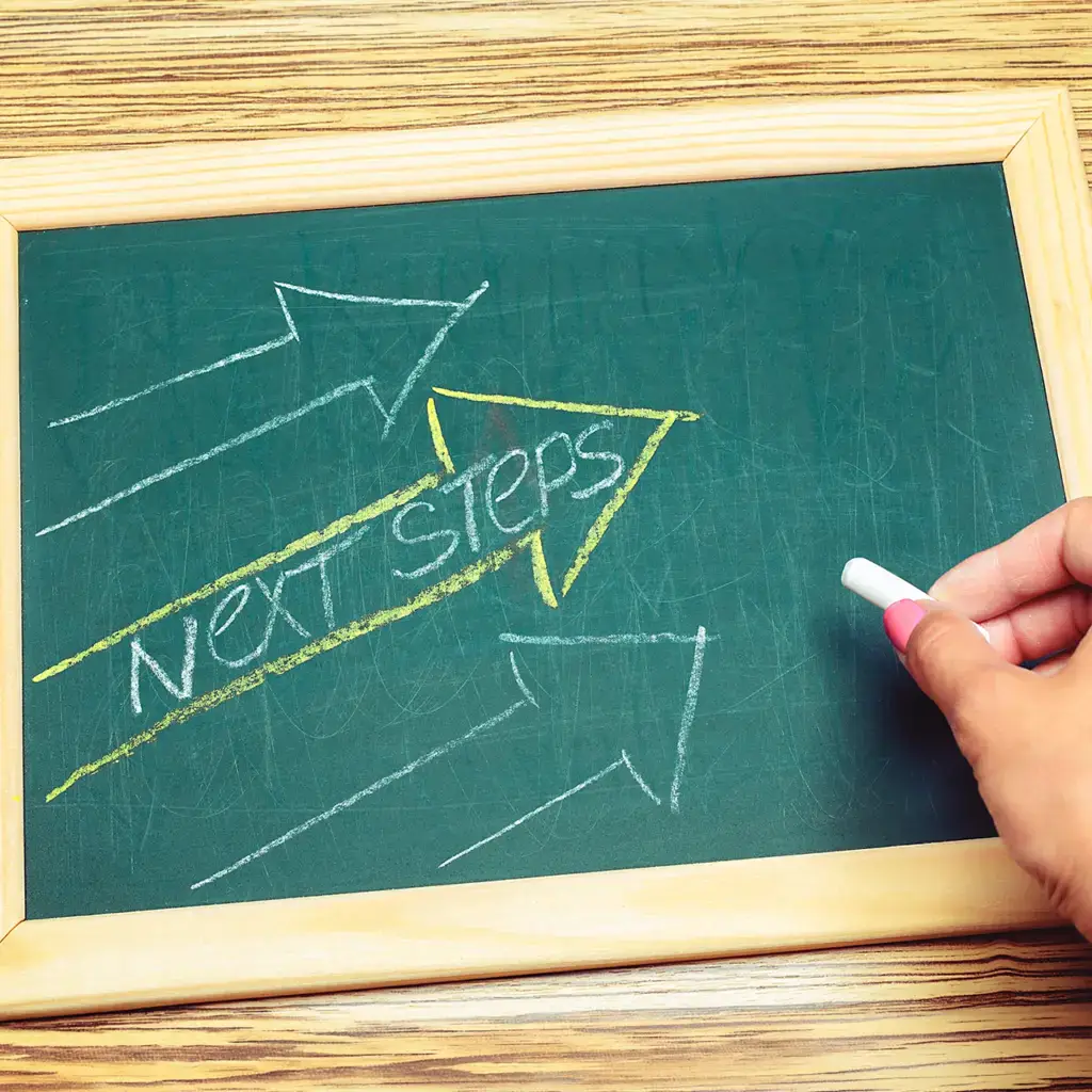 A hand writing 'NEXT STEPS' on a green chalkboard.
